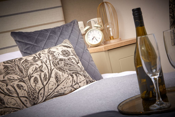 A bed with cushions, alarm clock and bottle of wine with glasses inside a Brickyard Lakes holiday lodge in North Yorkshire